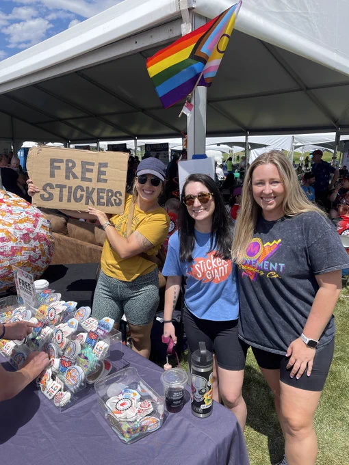 Three individuals at a festival booth under a tent, smiling and offering free stickers to attendees, with a rainbow flag visible in the background, promoting an inclusive atmosphere.