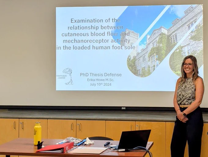 A photo of Erika at her defence, in front of a projected title screen with the same title of her thesis that is listed in this posting.