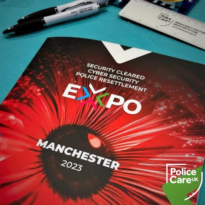 Image of a red Security Cleared Cyber Security Police Resettlement Expo 2023 brochure on a Police Care UK promotional table surrounded by Police Care UK pens and rulers. The white Police Care UK shield logo is in the bottom right corner