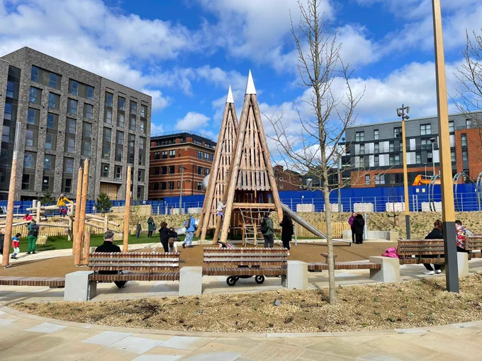 Pounds Park sits underneath a blue sky with white clouds, the park has two triangle shaped climbing frames with a row of benches in front and trees dotted about the site.