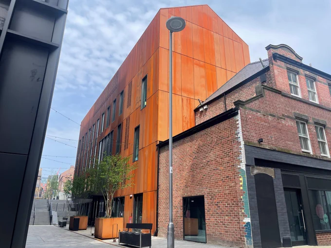 The building shown in the picture is Cambridge Street Collective, it has a rust coloured exterior with trees in front, the building next to it, in the foreground is made of bricks.