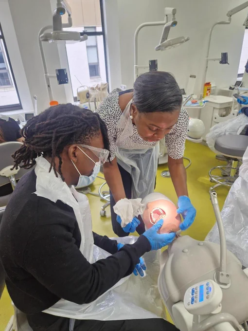 A student wearing a mask, apron and goggles is looking in to the mouth of a dummy with assistance from a teacher.