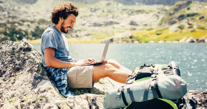 A man sitting outside in a scenic landscape while working on his laptop computer.