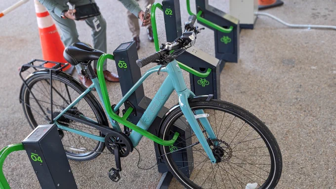 Alt Text: A blue and green bicycle docked at a public bike-sharing station, featuring multiple other bicycles and visible docking ports marked with numbers and green circular logos.