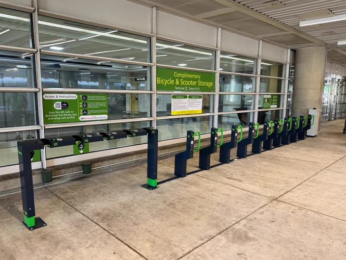 Row of empty bicycle parking racks at Reagan National Airport displaying signs reading "Complimentary Bicycle & Scooter Storage", emphasizing security and weather protection. A green and yellow sign above offers additional information on access and registration.