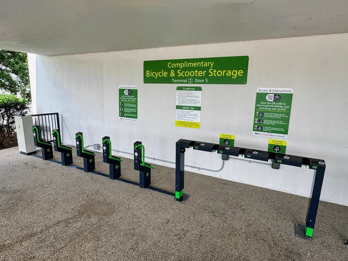 This image shows an area designated for complimentary bicycle and scooter storage at a facility, near Reagan National Airport Terminal 1 Door 5, as indicated by the signage. There are several empty bike racks lined up against a clear background. Three green signs are installed high on a wall, detailing usage policies, rules, and location information, emphasizing the complimentary aspect for both bicycles and scooters. The parking floor is a smooth grey, and the area is sheltered by a white overhead structure.