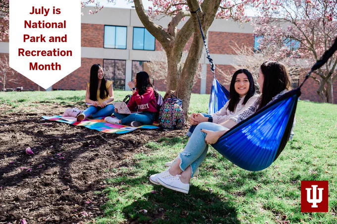 Two young women with long dark hair sit together in a blue hammock strung between two trees. In the background, two young women with long, dark hair sit on a blanket on the grass. Text on photo reads: "July is National Park and Recreation Month."