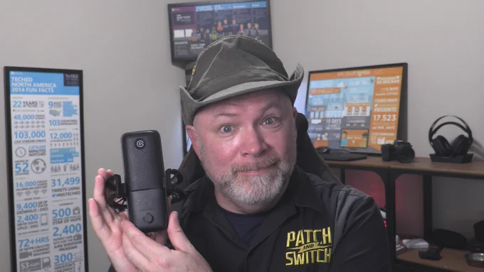 Rick Claus wearing Green hat, seated in podcasting studio, holding an Elgato Wave 3 USB mic - smiling into camera. 
