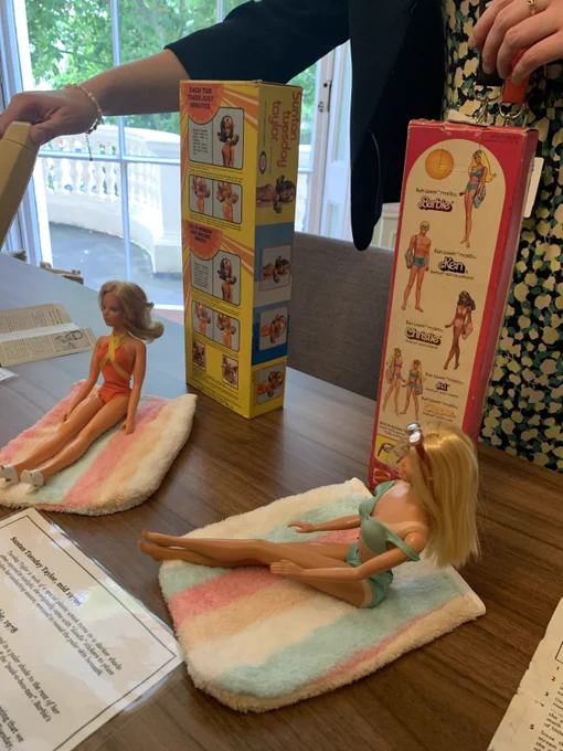 Two barbies are displayed on a wooden table, bearing visible tan lines 