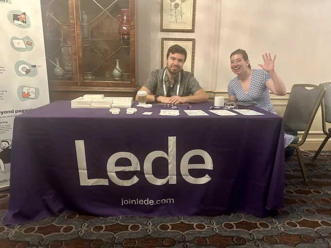 Lede CEO Matt Johnson and Lede principal software lead Christina Deemer sit at a conference booth looking at the camera. Christina is waving at the camera with her left hand. The table is covered in a purple linen with that says Lede in white letters and Lede's website address, joinlede.com. 