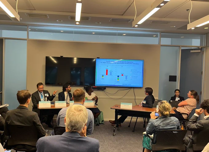 GCC's James Bradbury sits at a table with fellow panelists before an audience, gesturing at a slide showing graphics from GCC's analysis on emissions scenarios and federal infrastructure investments.