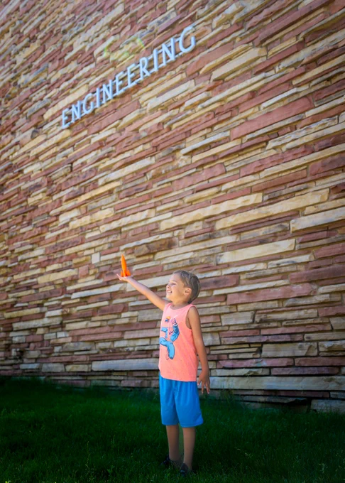 Image of a kid holding a 3-D printed rocket underneath the "Engineering" on the Engineering building at CSU. 