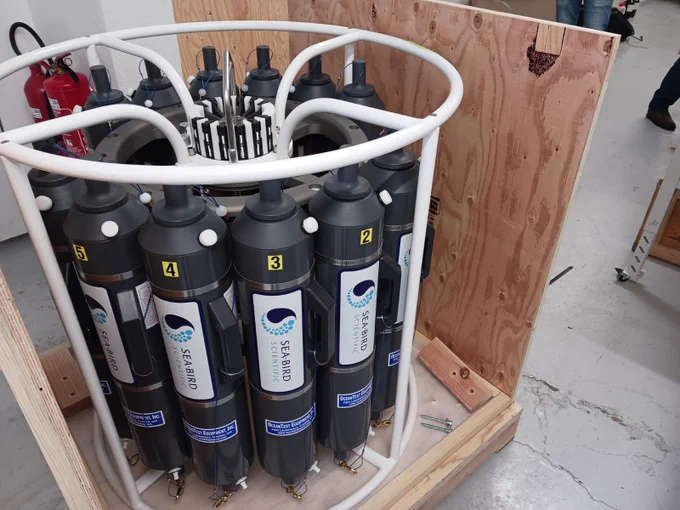 Equipment from Sea-Bird Scientific in a wooden crate, with 12 cylindrical sampling devices inside a white frame. Fire extinguisher and part of a person visible in the background.