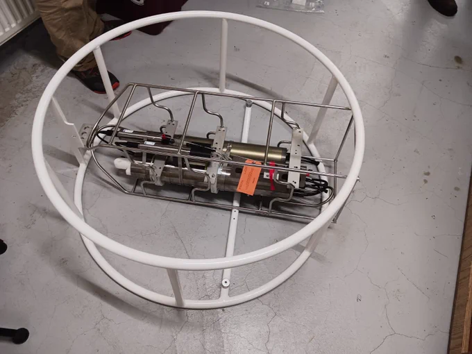 Cylindrical electronic device encased in a white metal cage lies on a gray concrete floor. Partial views of people's legs are visible nearby.