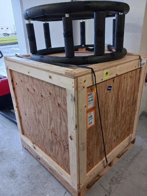 A large wooden crate and a cage with protective foam tubing around the top, situated indoors. Shipping labels and barcodes are attached to the crate. In the background, a forklift and an outdoor area are partially visible.