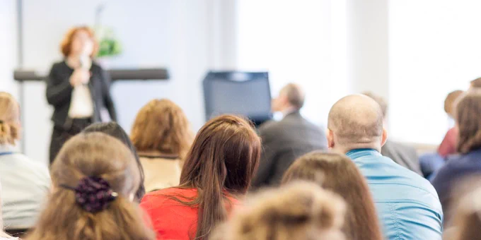 A presenter speaks at the front of a conference room filled with attendees facing forward, listening attentively.