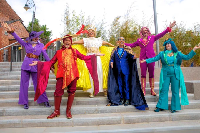 A group of six people in colourful, flamboyant costumes pose joyfully on outdoor steps. The costumes vary in design, incorporating bright hues like red, purple, yellow, and blue, and elaborate details such as ruffles, capes, and hats. The background features a brick building and a clear sky.