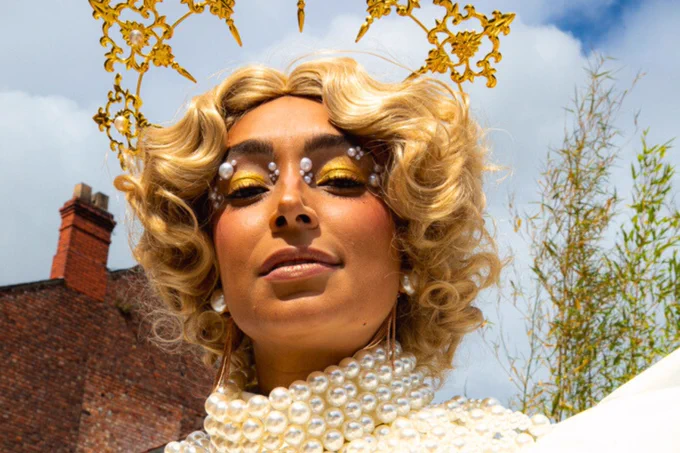 Person wearing a pearl necklace and a golden curly wig with decorative golden accents, posing under a clear blue sky.