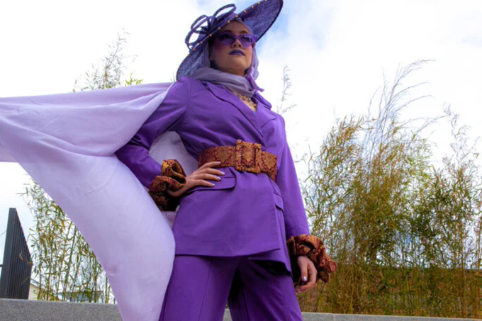 A person dressed in a vibrant purple witch costume with a hat and cape, poses confidently outdoors against a backdrop of tall grass and a clear sky.