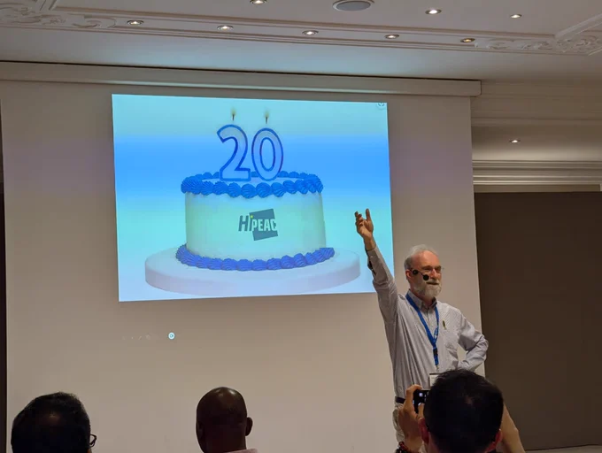 Koen De Bosschere standing in front of a slide featuring a blue-and-white cake with the HiPEAC logo and a 20 candle on top
