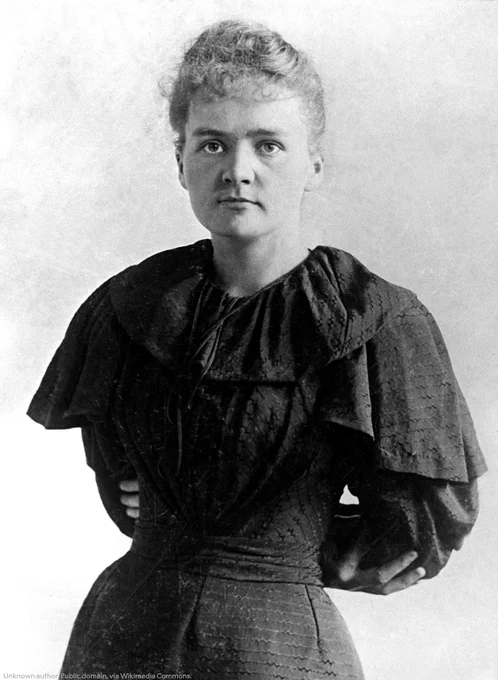 Portrait of Marie Curie, wearing a dark, high-collared dress with ruffled detailing. She appears in a formal pose, facing the camera with a composed expression.