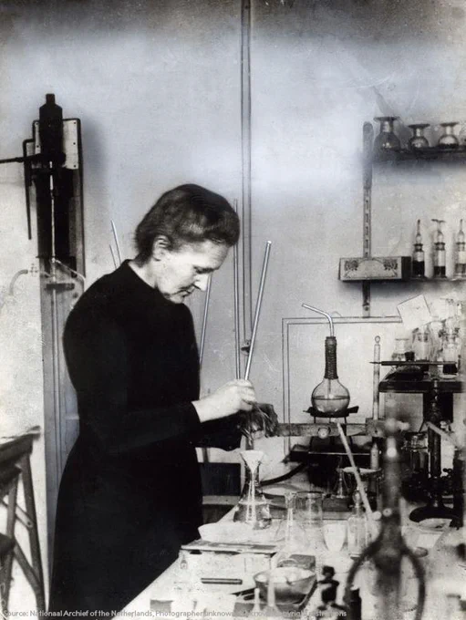Black and white historical photograph of Marie Curie in a laboratory. She is engaged in an experiment, using a pipette and a flask. The background is filled with various scientific glassware and equipment. 