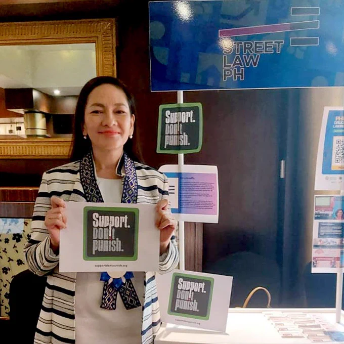 Senator Risa Hontiveros, a lady from the Philippines with straight black hair, round bright metal earrings, and a striped jacket, holds up the Support. Don't Punish logo next to a StreetLawPH stand at the Philippine Drug Summit.