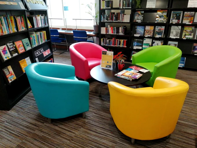 Four comfy, soft-covered chairs - 1 pink, 1 yellow, 1 light green, 1 light blue - placed in a circle around a low desk with bookshelves and a window behind.