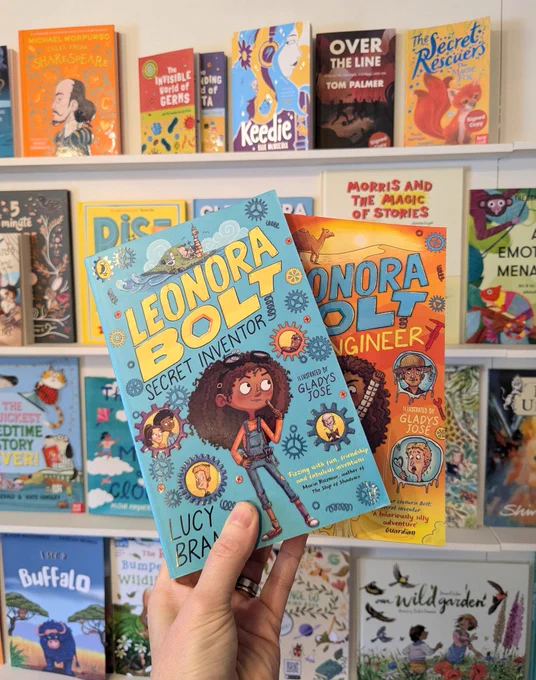 Holding two copies of Leonora Bolt Secret Inventor books in front of children's book display 