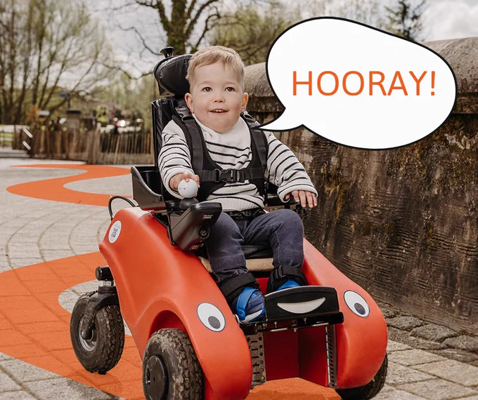 A young child using a Wizzybug powered wheelchair with a speech bubble saying 'Hooray!'