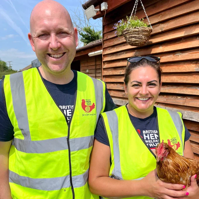 A man and a woman are in high-vis British Hen Welfare Trust vests, and the woman is holding a hen.
