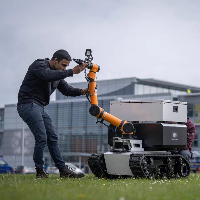 A grassy field with two men working on a robot. The robot has a long orange arm with camera equipment on the end. 