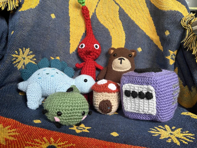 a selection of plushies grouped together on a blanket, including a junimo, gamecube, pikmin, mushroom house, bear, and dino. 