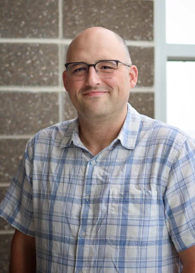 Dr. Micah Ferrell smiles at the camera. He is wearing a blue and white plaid shirt and glasses and is standing in front of a gray and white stone wall and a window.
