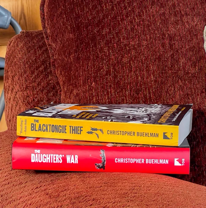 The Blacktongue Thief and The Daughters' War by Christopher Buehlman are stacked on a brown couch
