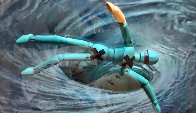 A wooden moveable art figurine painted green with a black cross on the hips and on the chest. The figurine is falling backwards into a dark grey-blue swirling whirlpool 