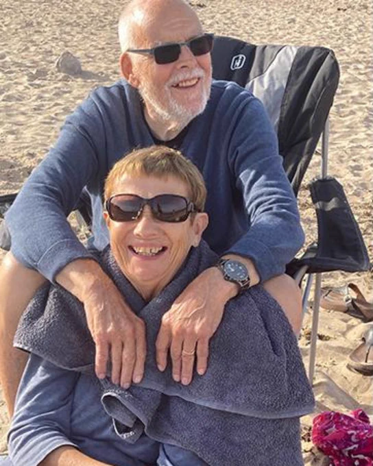 Ralph and Glenys are at the beach. Ralph is sat in a black chair with his arms resting on Glenys shoulder, who is front of him. They are both wearing sunglasses and both have a smile on their face. 