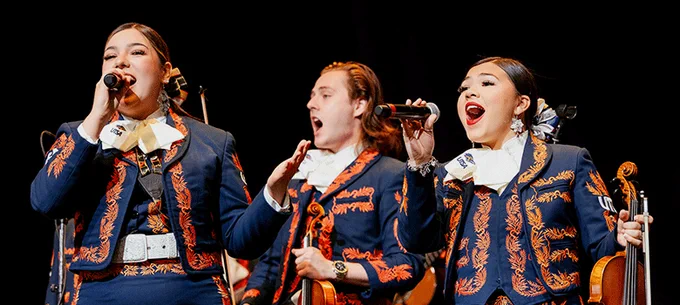 Three mariachi performers on stage singing into microphones, dressed in traditional charro outfits, with one holding a violin.