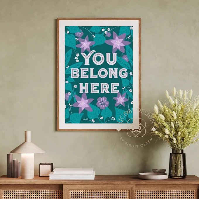 A framed poster with the text "You Belong Here" in bold letters. The background features a vibrant floral design with purple flowers and green leaves. The poster is displayed on a light green wall, above a wooden console table adorned with books, a modern lamp, and a vase of dried flowers. The design is by LiCoBi Design by Birgit Olzem.