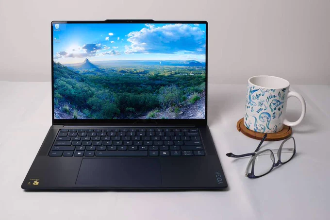Photo of Lenovo Yoga Slim 7x Snapdragon laptop open on a desk with a mug and a pair of glasses to its right.