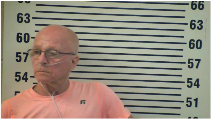 Defendant standing in front of backdrop for arrest photo with oxygen tube.
