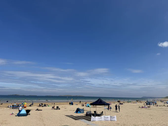A bright blue sky with some brief clouds, above a sandy beach with beach users and a gazebo and windbreakers with the Mae Murray Foundation logo