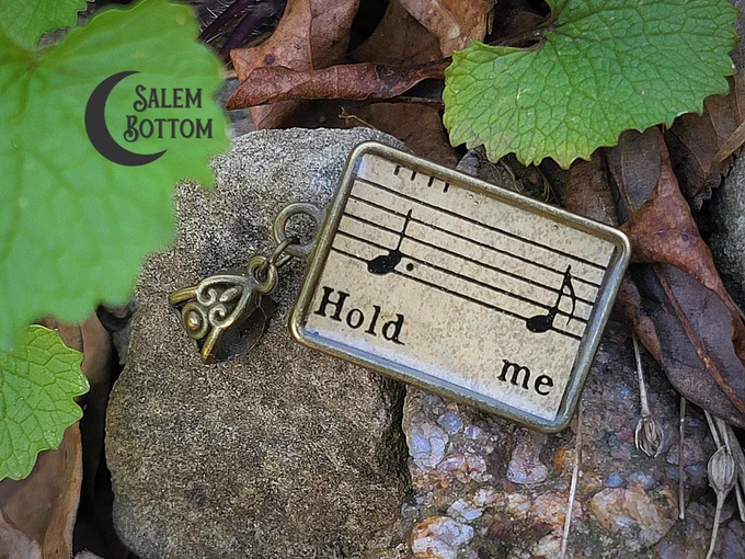 Antique sheet music pendant - bronze in color - with the words "Hold me" and two music notes, set atop a gray rock near some green and brown leaves