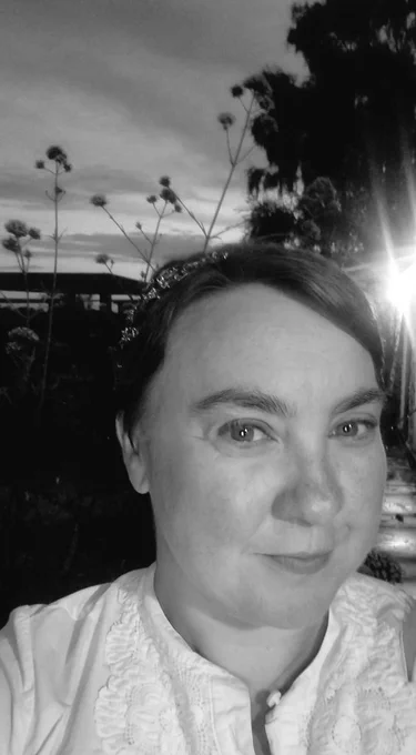 Stephe smiles to the camera. She wears a white shirt, and a crown-like headband, shaped to look like branches and twigs. Behind her, a moody summer sky.