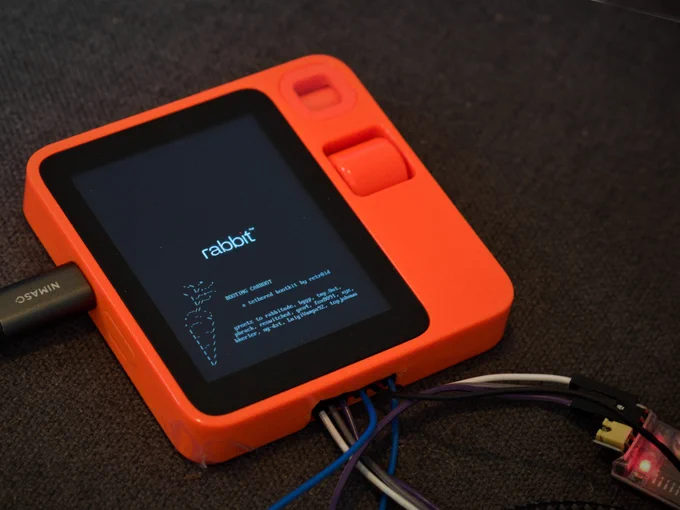 A rabbit R1 (a bright orange plastic square, with an LCD panel), with jumper wires coming out of a hole in the side, showing the 'carroot' boot screen, which includes an ascii art carrot