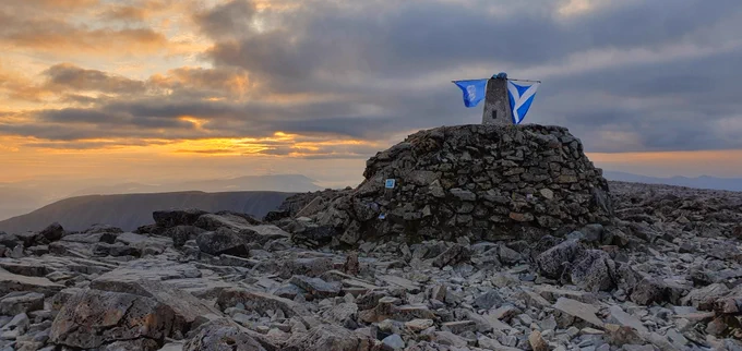 The sun rising over the summit of Ben Nevis with two flags flying from a cairn