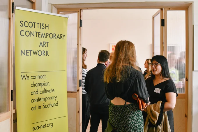 Colour photograph of some people chatting in a doorway with a yellow banner reading 'SCOTTISH CONTEMPORARY ART NETWORK' next to them.