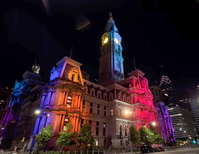 A large historic building illuminated with colorful lights, including blue, purple, and red, at night. Trees and a streetlight are visible in the foreground.