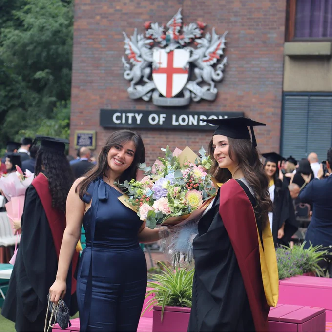 Two people standing in front of the City of London crest holding flowers, one is wearing a graduation cap and gown