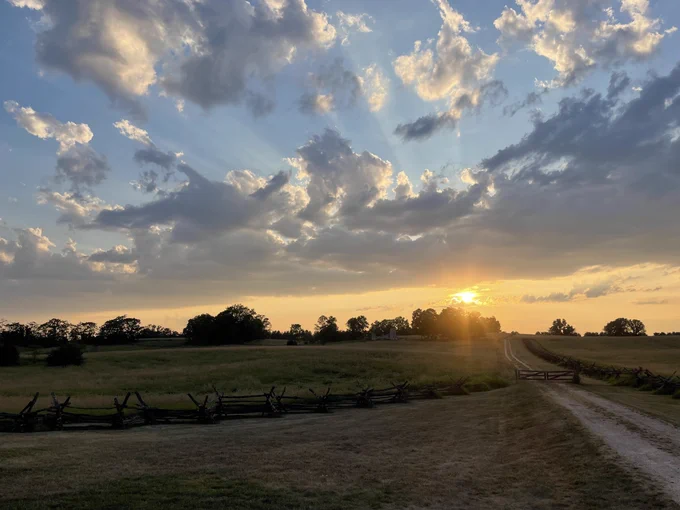 An orange and blue sky with a sunet above a above a green tree line and field.